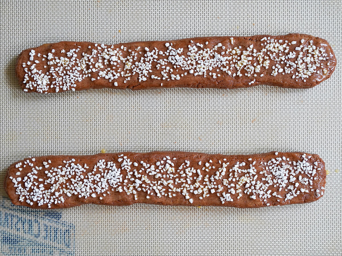 two logs of cookie mixture topped with pearl sugar sitting on silicone mat before baking.