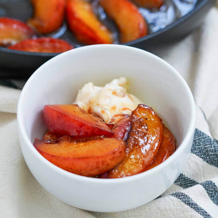 bowl of caramelized peaches with ice cream at back of bowl and skillet of peaches behind.