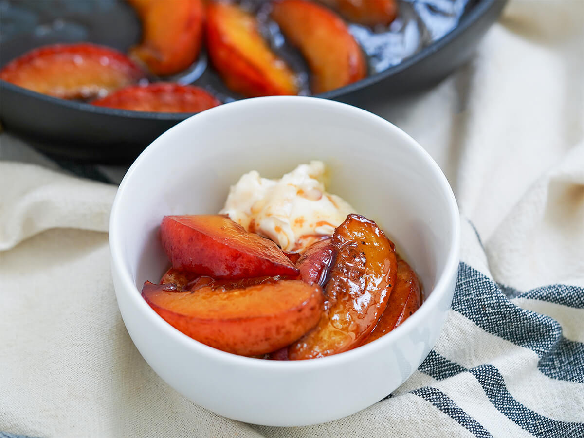 bowl with caramelized peaches with ice cream and skillet with more peach slices behind.