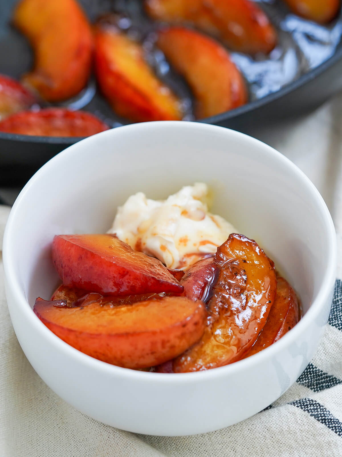 close view of bowl of caramelized peaches with ice cream and skillet of peaches behind.