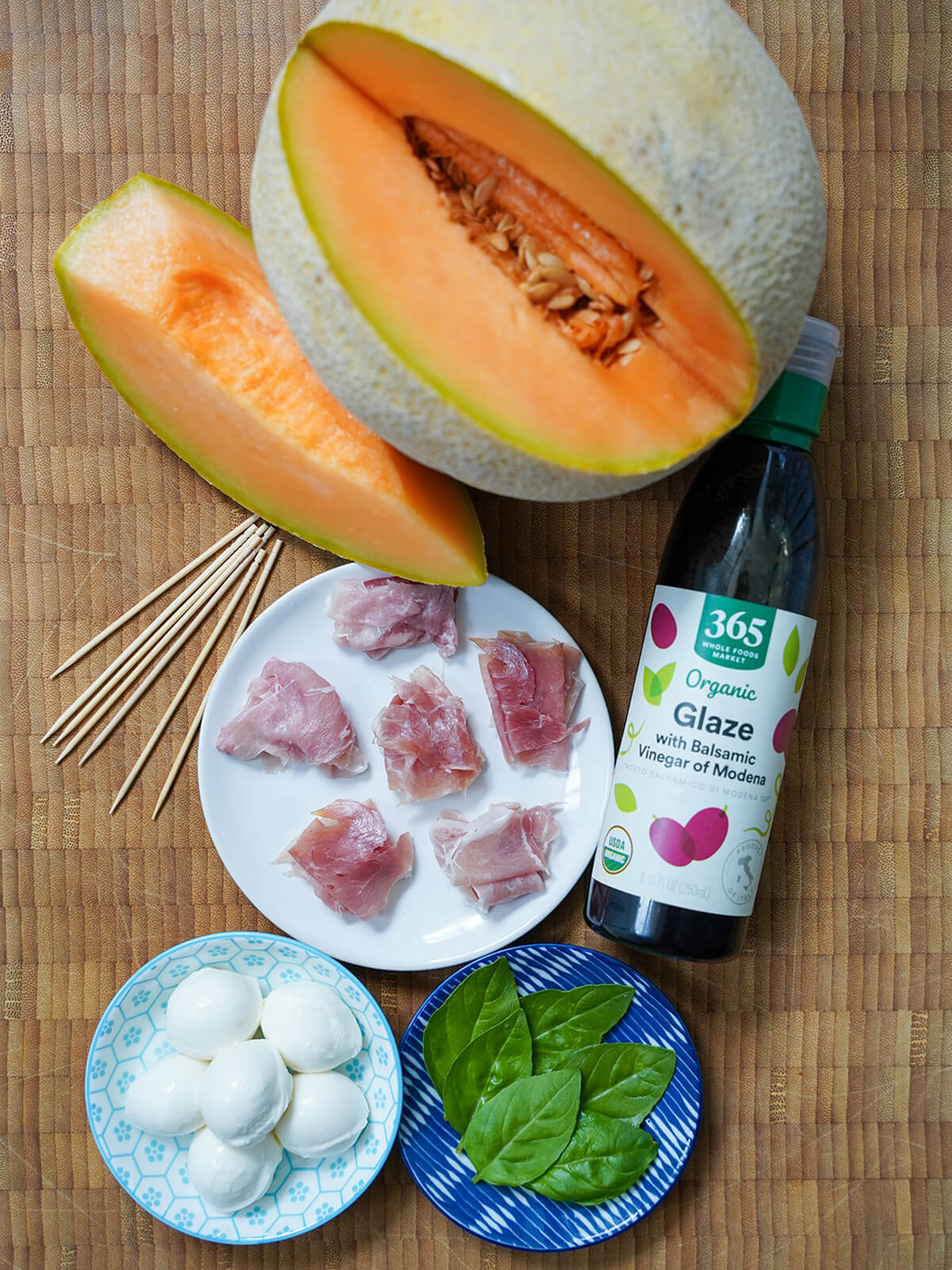 cantaloupe melon with a wedge cut from it, bottle of balsamic glaze, plate with stacks of prosciutto, small dishes with basil leaves and balls of mozzarella.