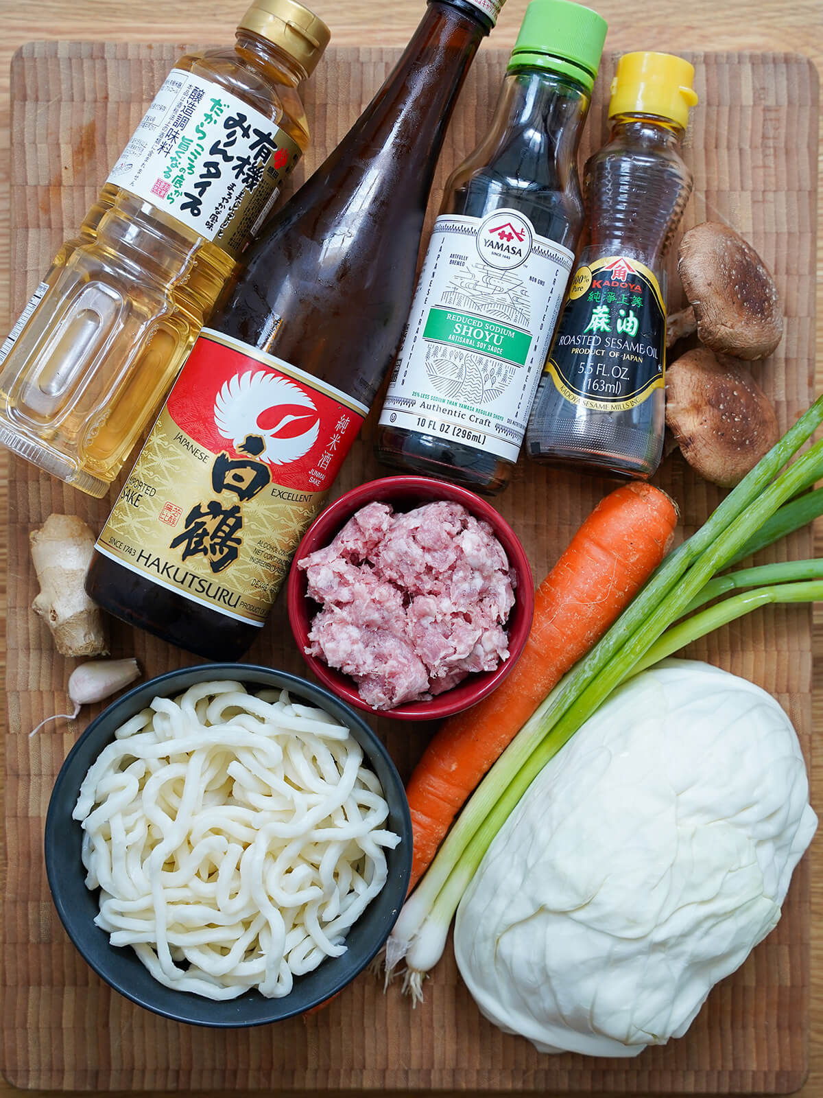 bottles of soy, sake, mirin and sesame oil with dishes with ground pork, udon noodles and carrot, cabbage, green onions and shitake mushrooms to side.