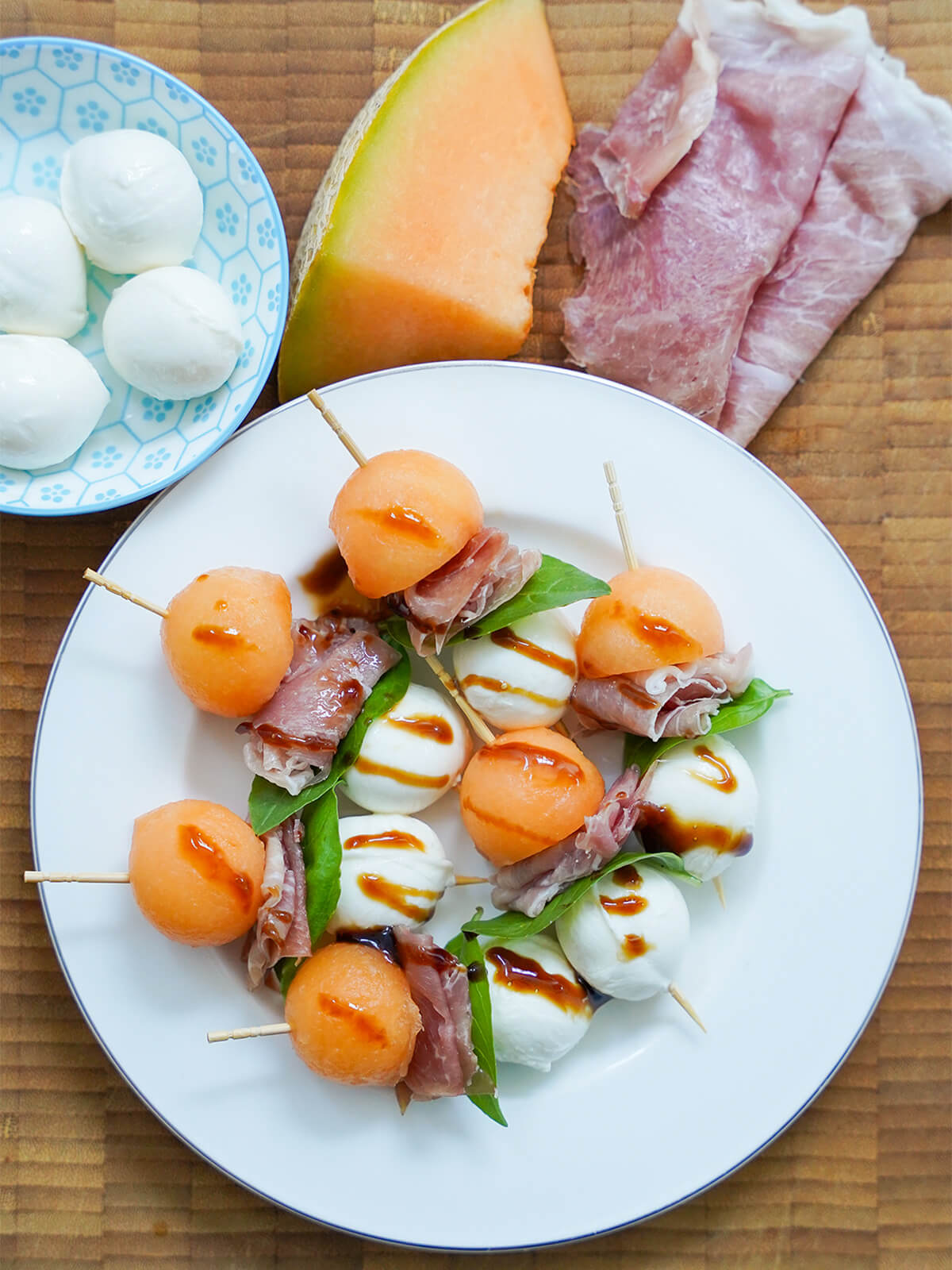 plate of melon and prosciutto skewers with chunk of cantaloupe, slices of ham and some mozzarella balls above plate.