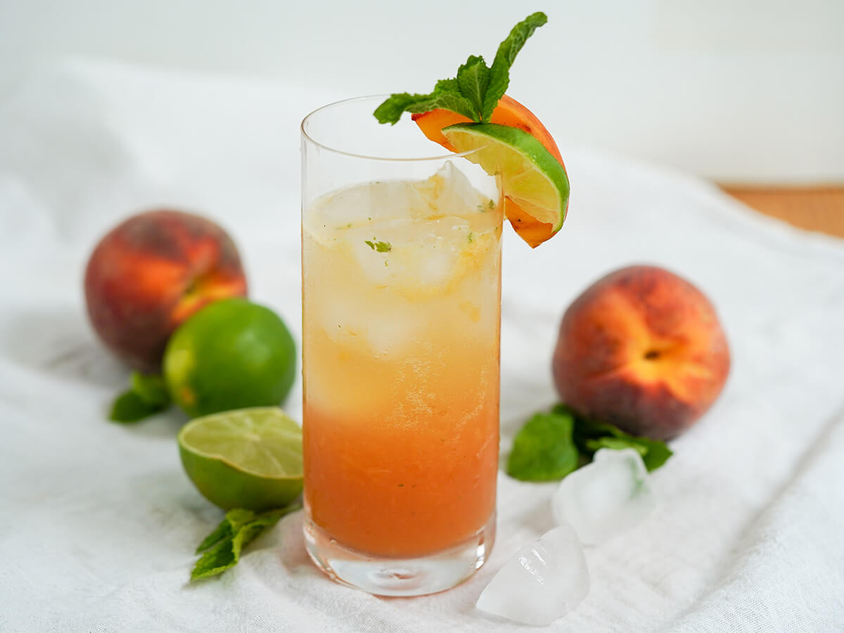 side view of glass of peach mojito with two peaches and a lime behind glass.
