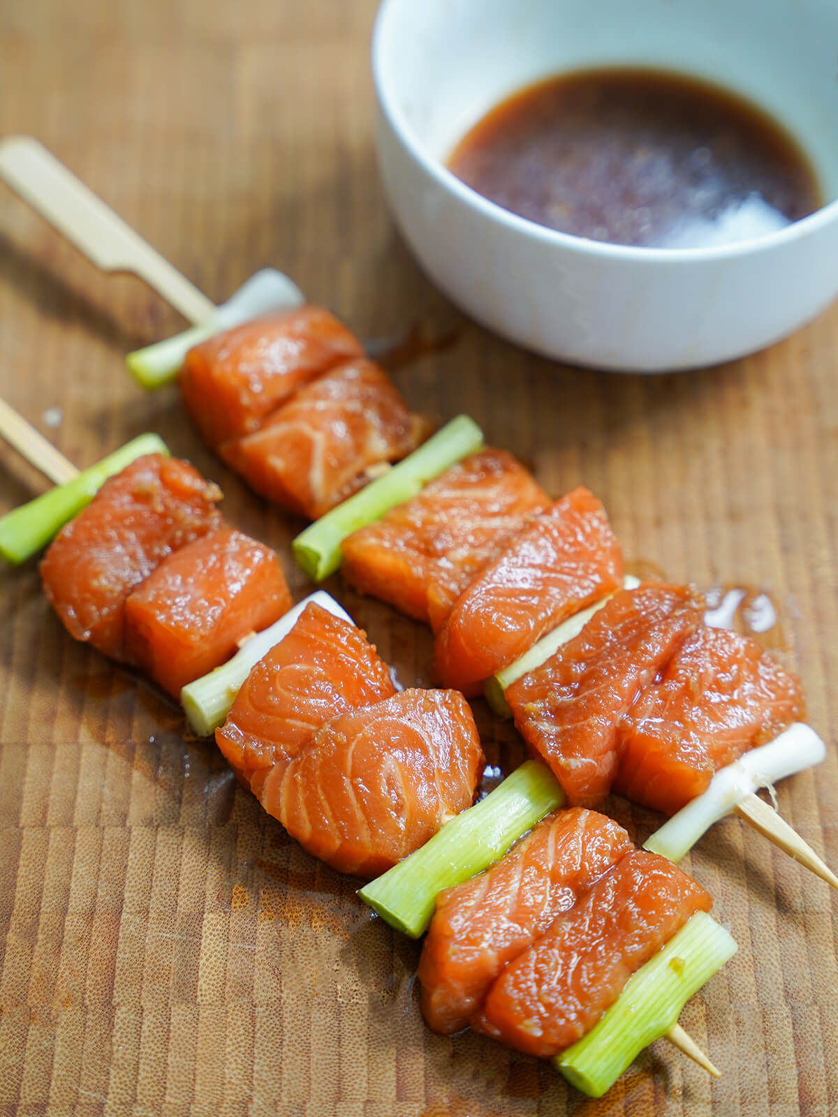 pieces of marinated fish and green onion on wooden skewers with bowl of leftover marinade behind.