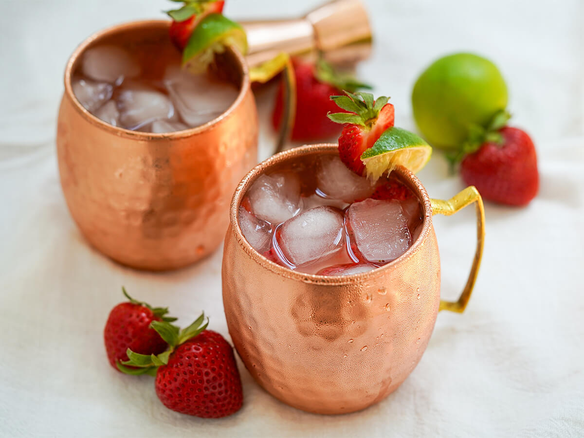 two copper mugs of strawberry Moscow mule with strawberries and lime to sides.
