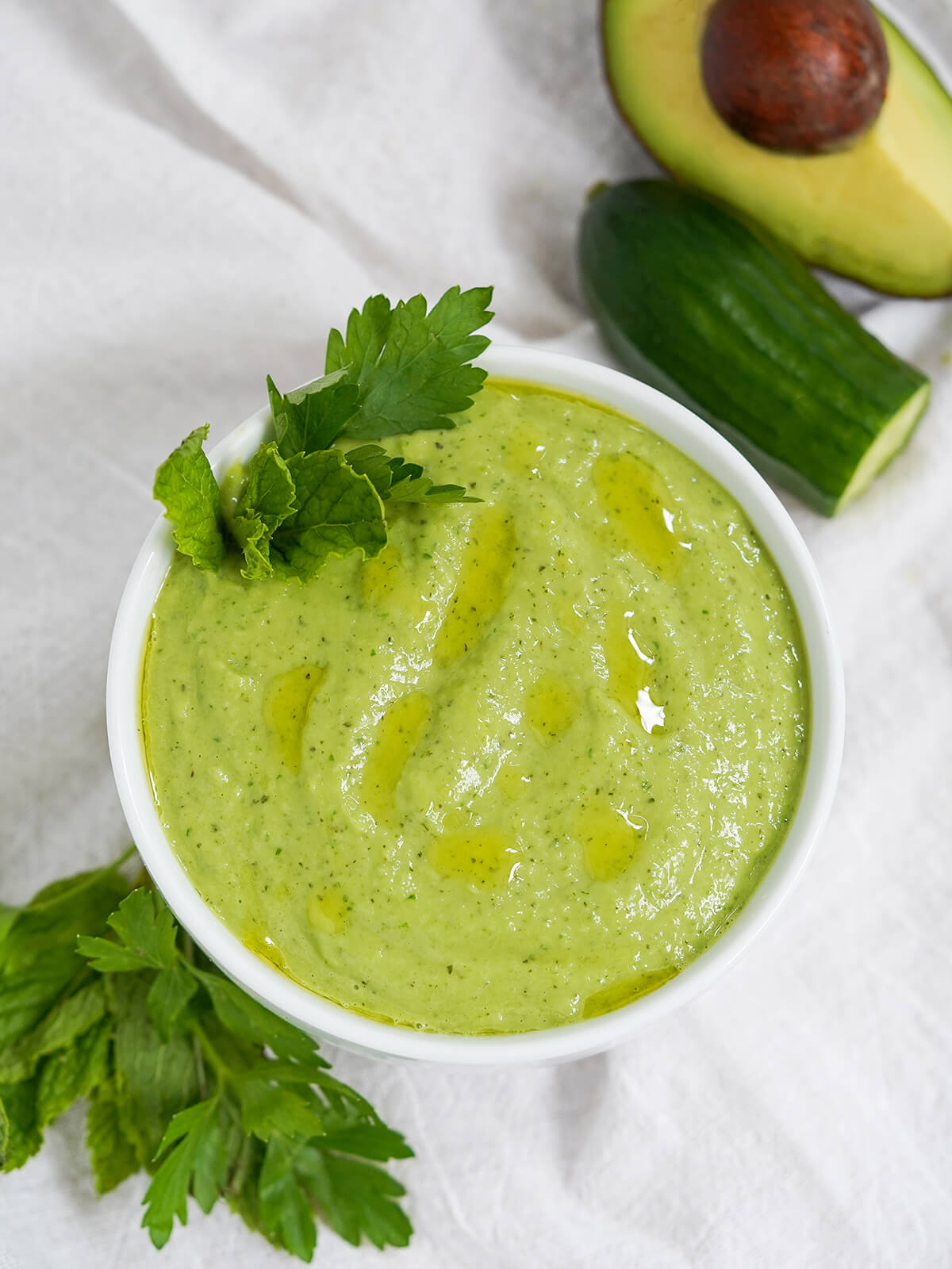 overhead view of bowl of cucumber avocado gazpacho (green gazpacho) with part cucumber and avocado above and bunch of herbs below bowl.