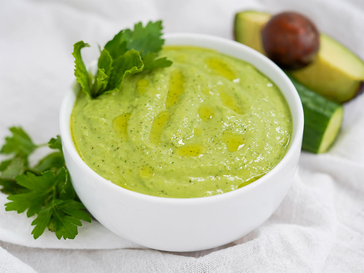 side view of bowl of cucumber avocado gazpacho (green gazpacho) with herbs to side of bowl.