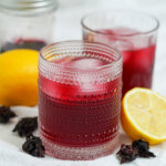 glass of hibiscus lemonade with lemons either side and glass in background.