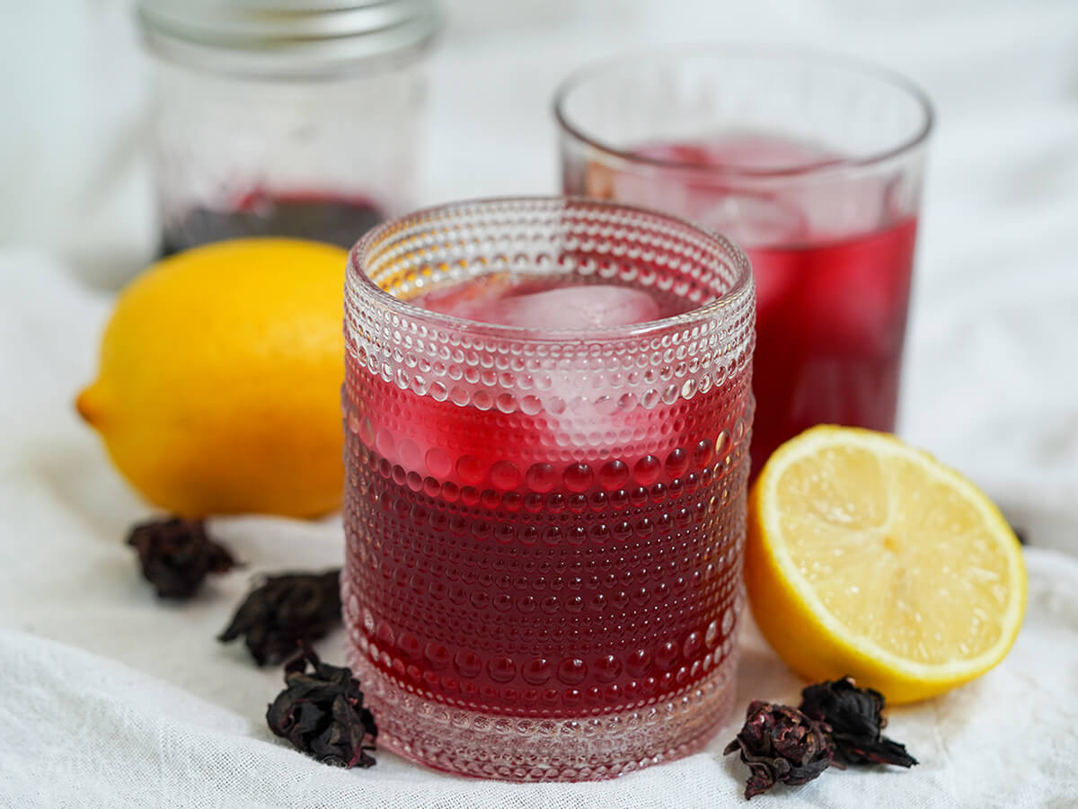 side view of two glasses of hibiscus lemonade, one of them behind other, with cut lemon one side, whole lemon behind and hibiscus flowers to side.