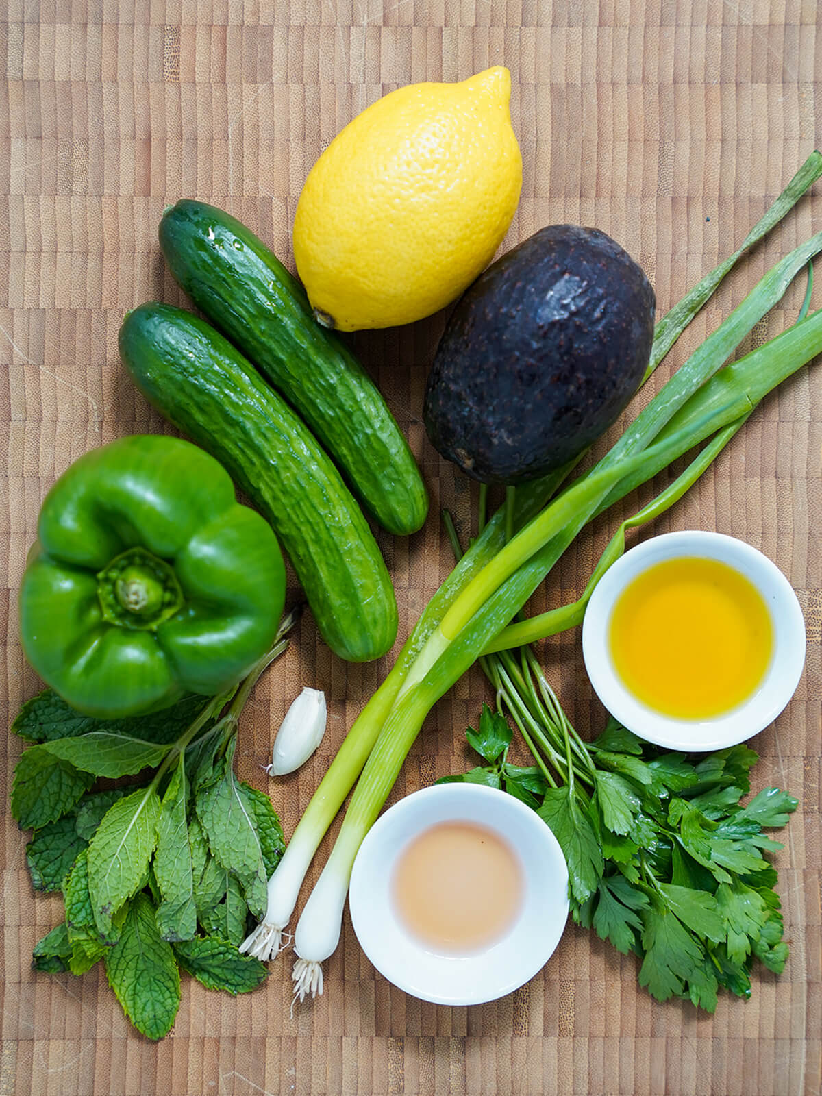mini cucumbers, green pepper, avocado, lemon, green onions, garlic, herbs and small dishes with vinegar and olive oil on wooden board from overhead.