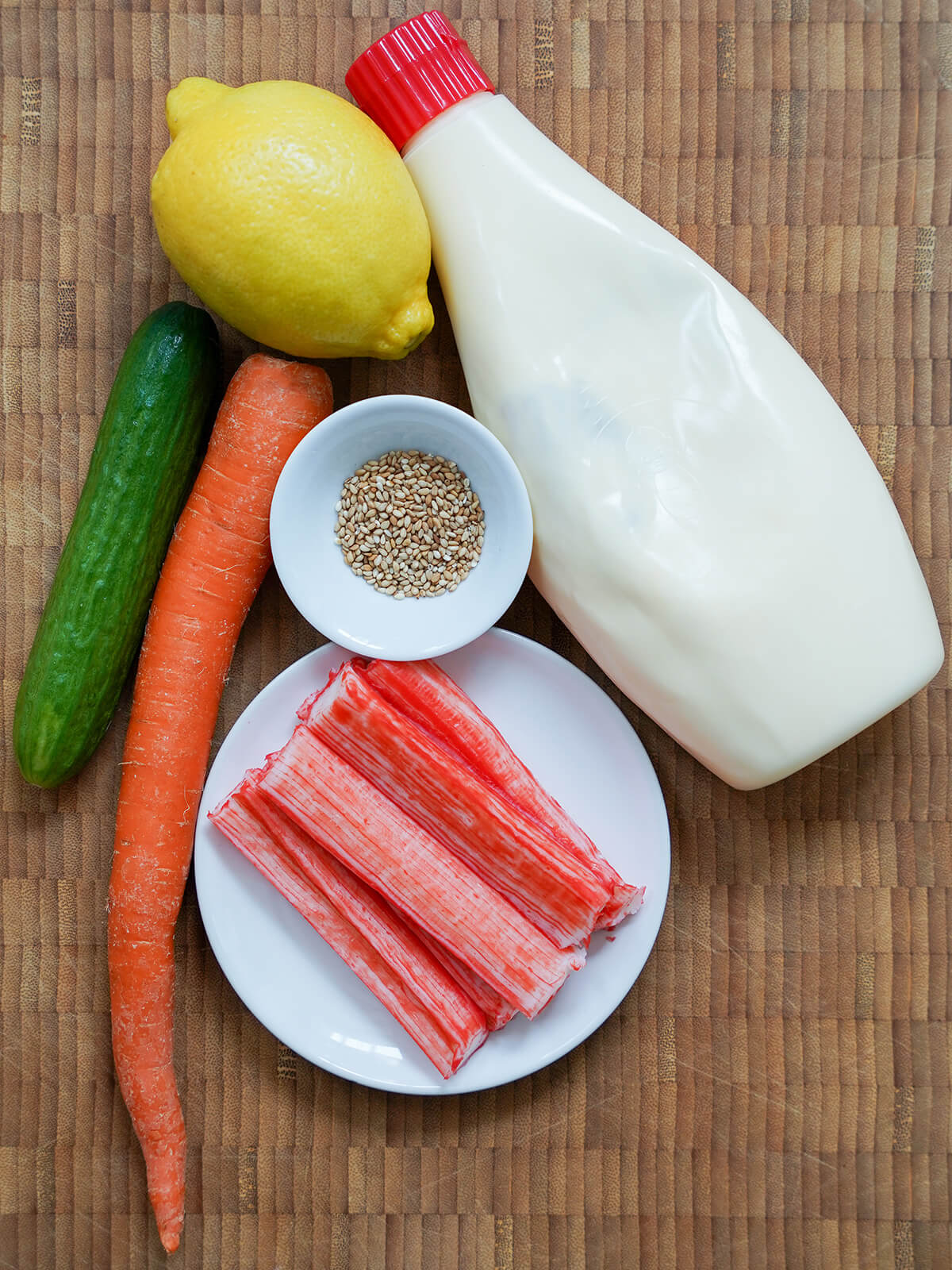 bottle of Japanese mayonnaise, lemon, bowl with sesame seeds, imitation crab sticks on plate and carrot and baby cucumber to side.