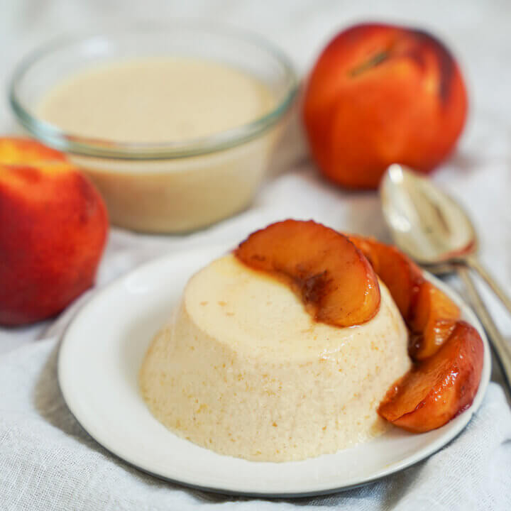 plate of peach panna cotta with caramelized peaches on top and to side with peaches in background behind plate.