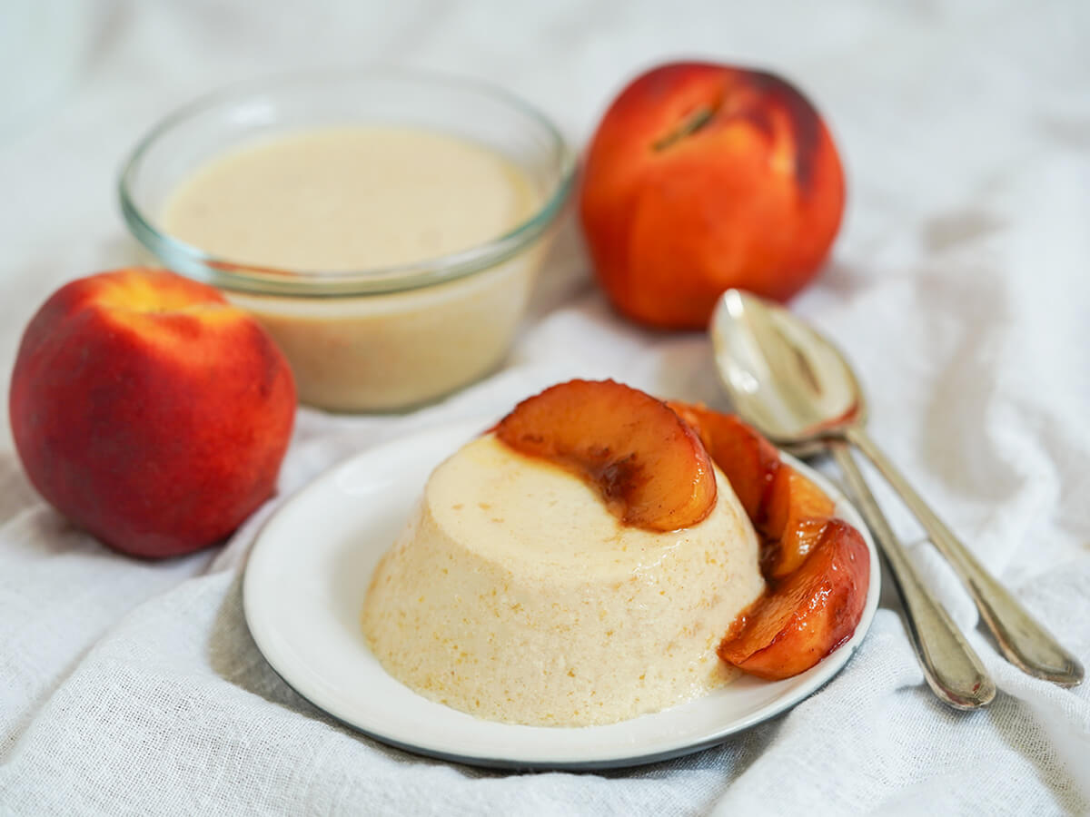 plate of peach panna cotta topped with peach slices with another in dish behind, peaches and spoons to side.