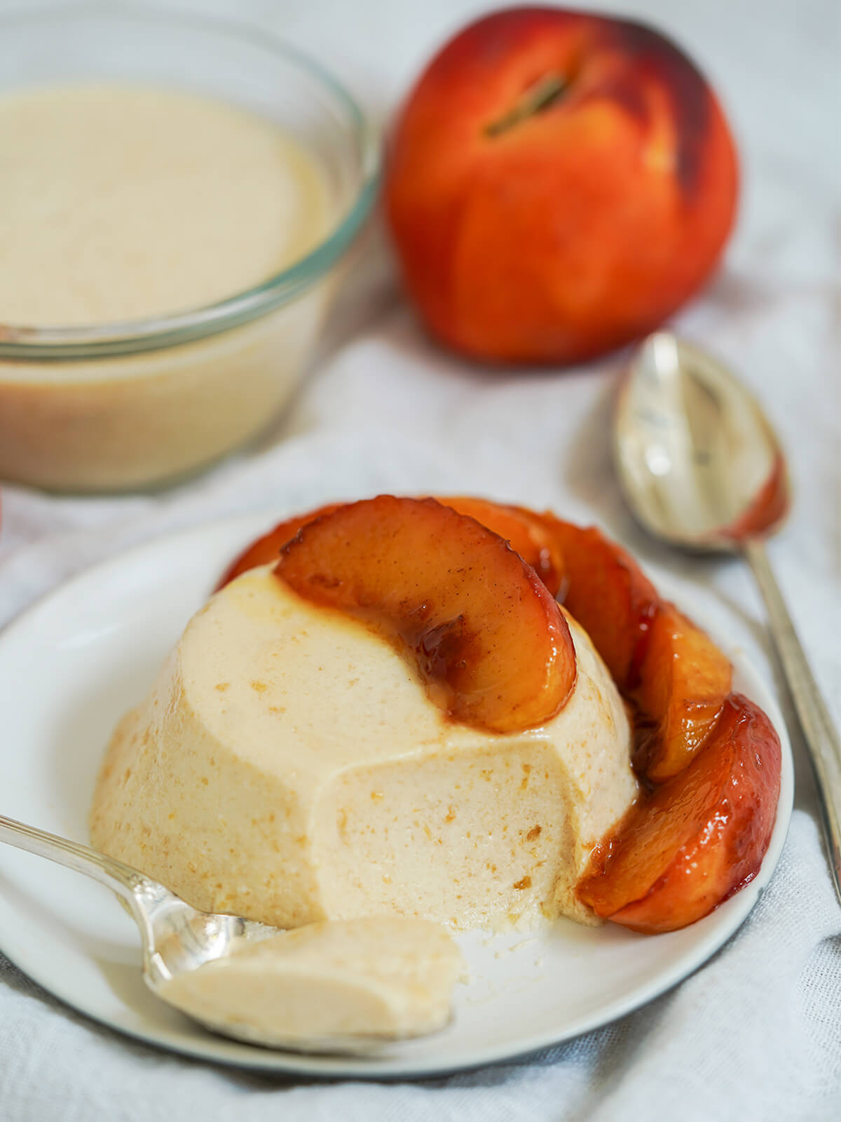 peach panna cotta on dish topped with caramelized peach slices with a spoonful taken out and sitting to side.