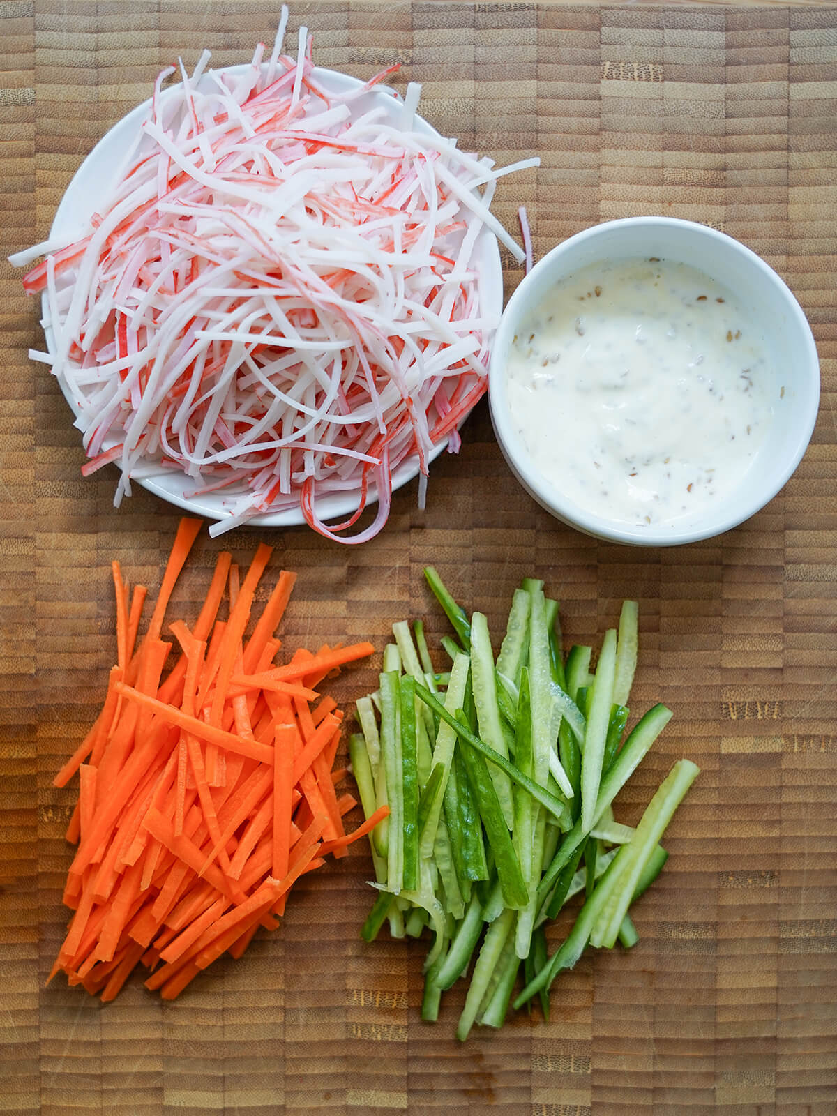 plate of shredded imitation crab, bowl of mayo-based dressing and piles of julienned carrots and cucumber on wooden board.