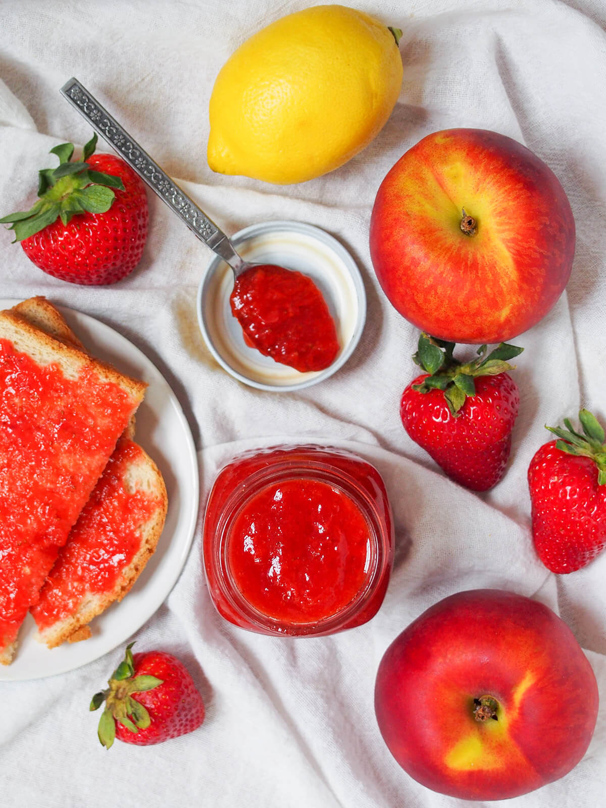 overhead view of jar of strawberry peach jam with peaches, strawberries and lemon around, toast with jam to side and spoon of jam resting on lid.