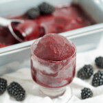 glass of blackberry sorbet with blackberries to either side of glass and dish of sorbet blurred in background.