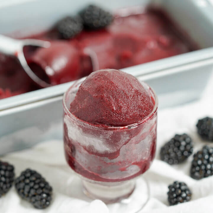 glass of blackberry sorbet with blackberries to either side of glass and dish of sorbet blurred in background.