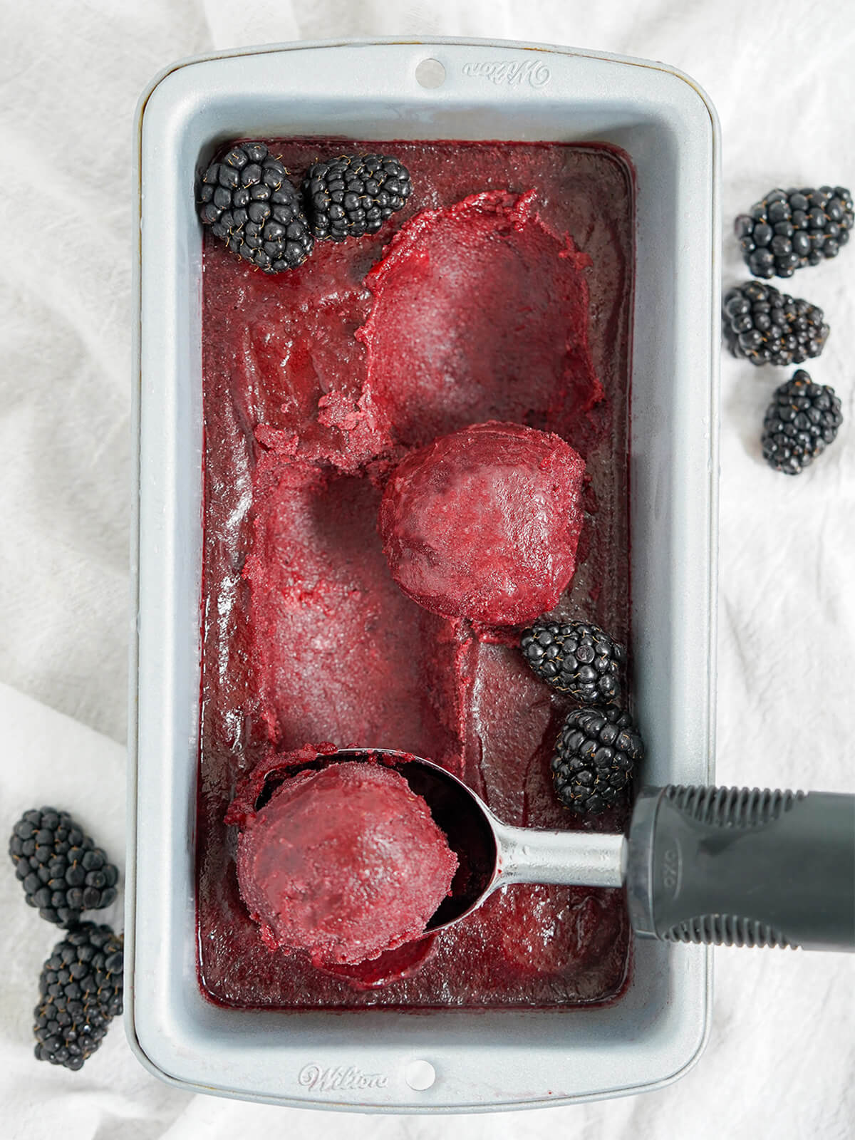 loaf pan filled with blackberry sorbet with scoops sitting on top and in ice cream scoop.