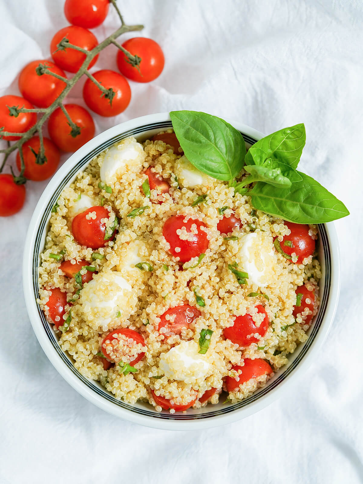 This Caprese quinoa salad is a delicious, light & healthy salad that's perfect for a picnic, potluck or as a side to many a meal. It's easy to make, versatile and easy to add to, as well. overhead view of bowl of caprese quinoa salad with cherry tomatoes on vine above one corner.
