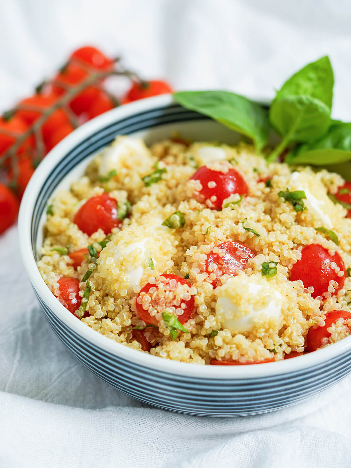 This Caprese quinoa salad is a delicious, light & healthy salad that's perfect for a picnic, potluck or as a side to many a meal. It's easy to make, versatile and easy to add to, as well. side part view of bowl of caprese quinoa salad with tomatoes on vine part in view behind.