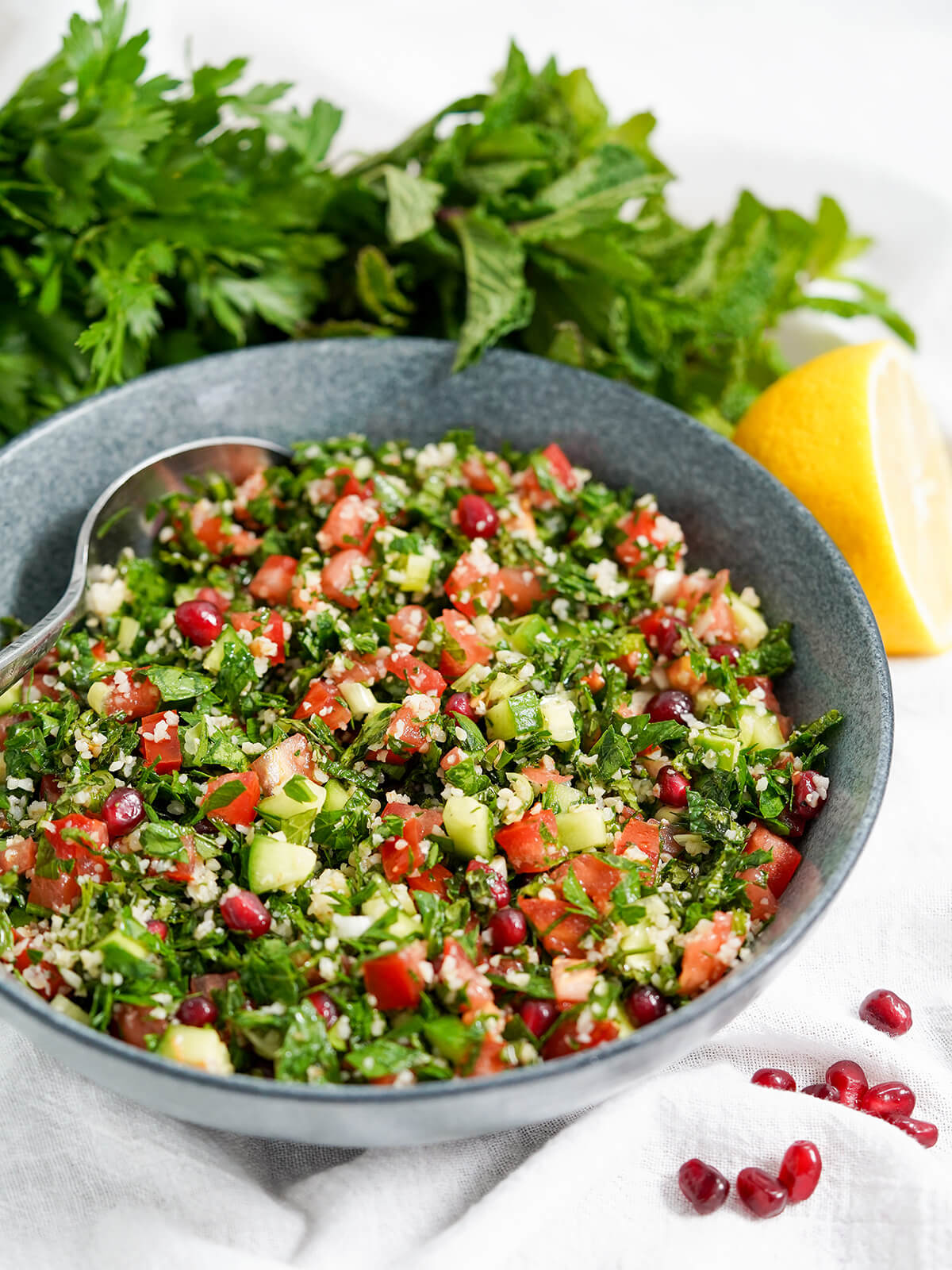 This tabbouleh with pomegranate is wonderfully fresh, bright and packed with flavor. It's easy to make and a delicious side to many a meal. side view of bowl of tabbouleh with pomegranate with bunches of herbs behind.
