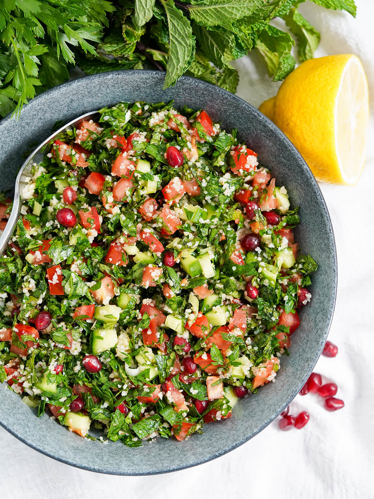 tabbouleh with pomegranate is a wonderfully fresh and tasty Middle Eastern salad overhead view of bowl of tabbouleh with pomegranate with lemon to side and herbs above.