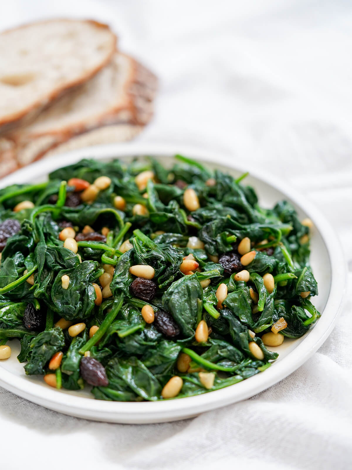 plate of Catalan spinach with pine nuts and raisins from side with bread blurred in background.