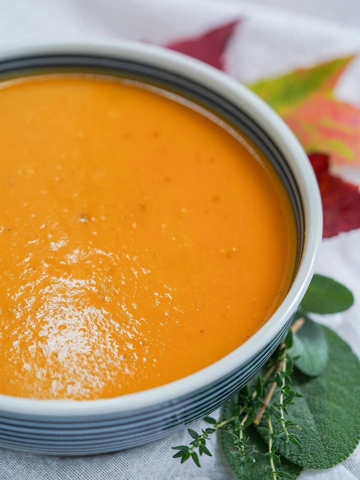 This butternut squash soup with apple is such a warming bowl of fall flavors. The flavors are that bit more complex than your average bowl, with the sweetness of the squash balanced out by a gently savory note from sage and thyme. side partial view of bowl of butternut squash apple soup with herbs to side of bowl.