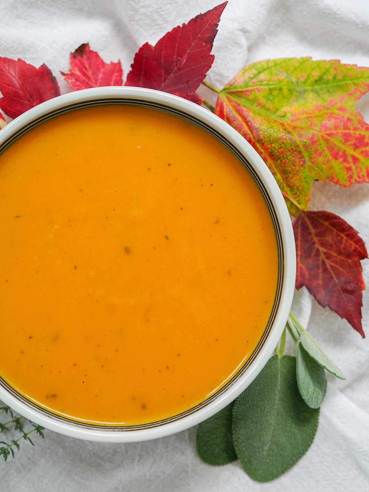 This butternut squash soup with apple is such a warming bowl of fall flavors. The flavors are that bit more complex than your average bowl, with the sweetness of the squash balanced out by a gently savory note from sage and thyme. overhead view of bowl of butternut squash apple soup with leaves to side of bowl.