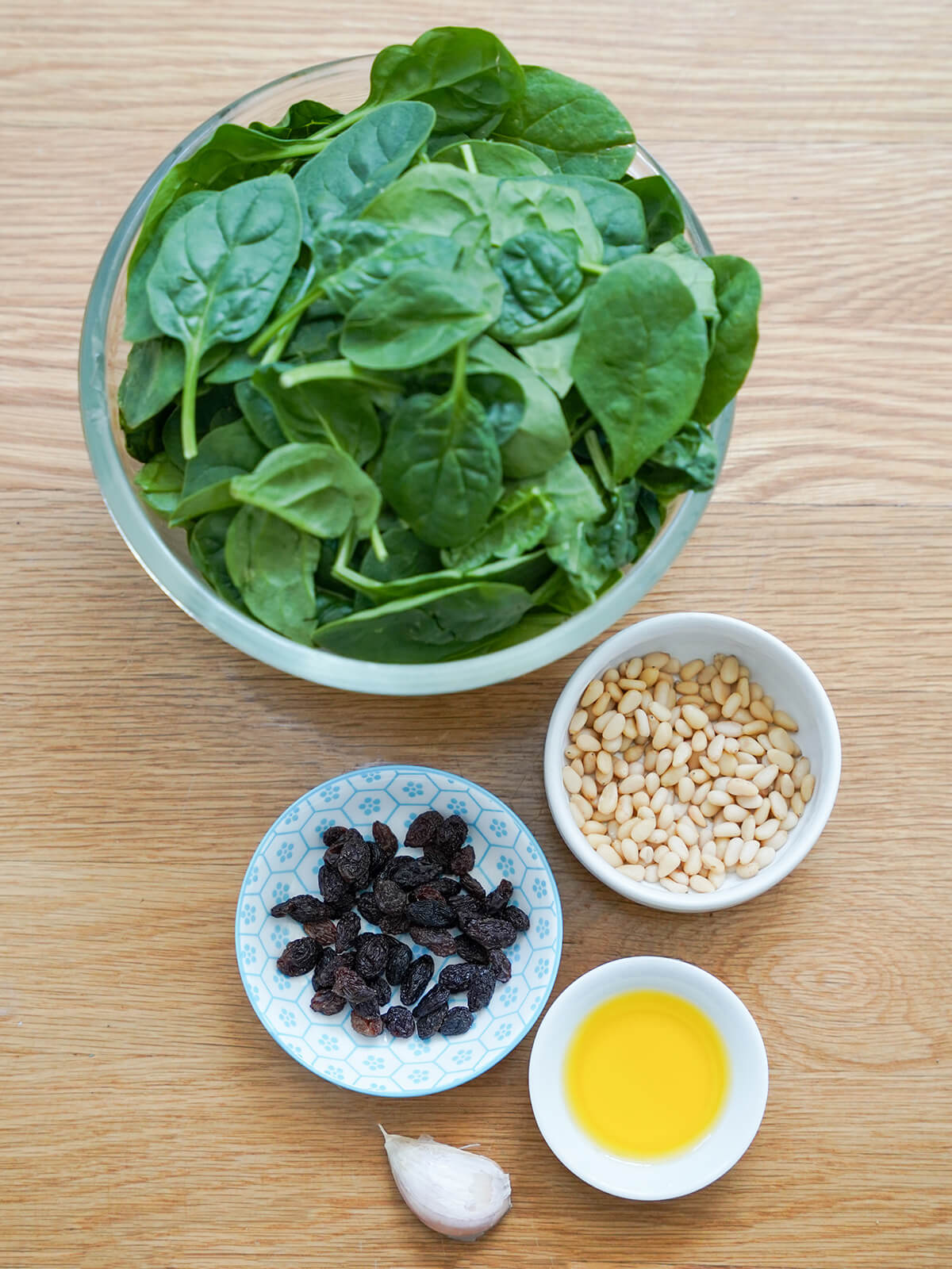 large bowl of spinach with dishes of raisins, pine nuts, olive oil and a garlic clove below.