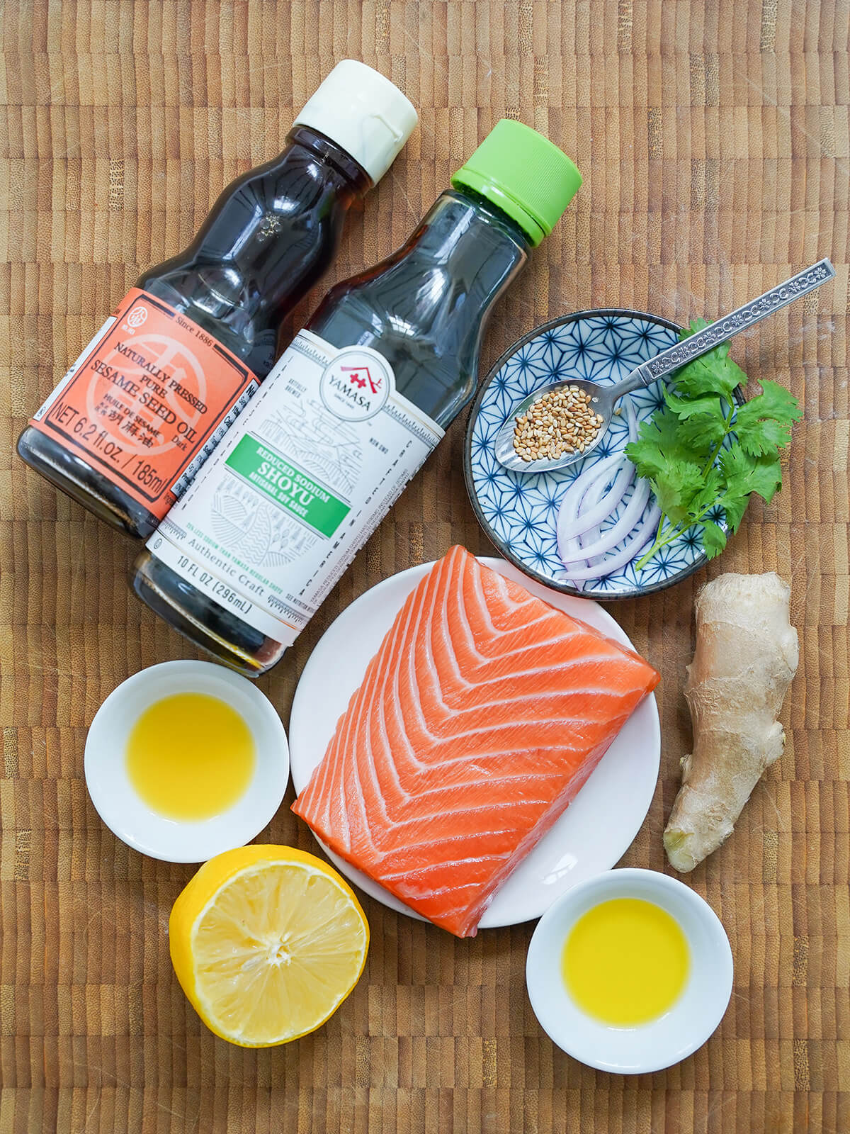 piece of salmon surrounded by small dishes with oil and juice, lemon, ginger and bottles of soy and sesame oil.