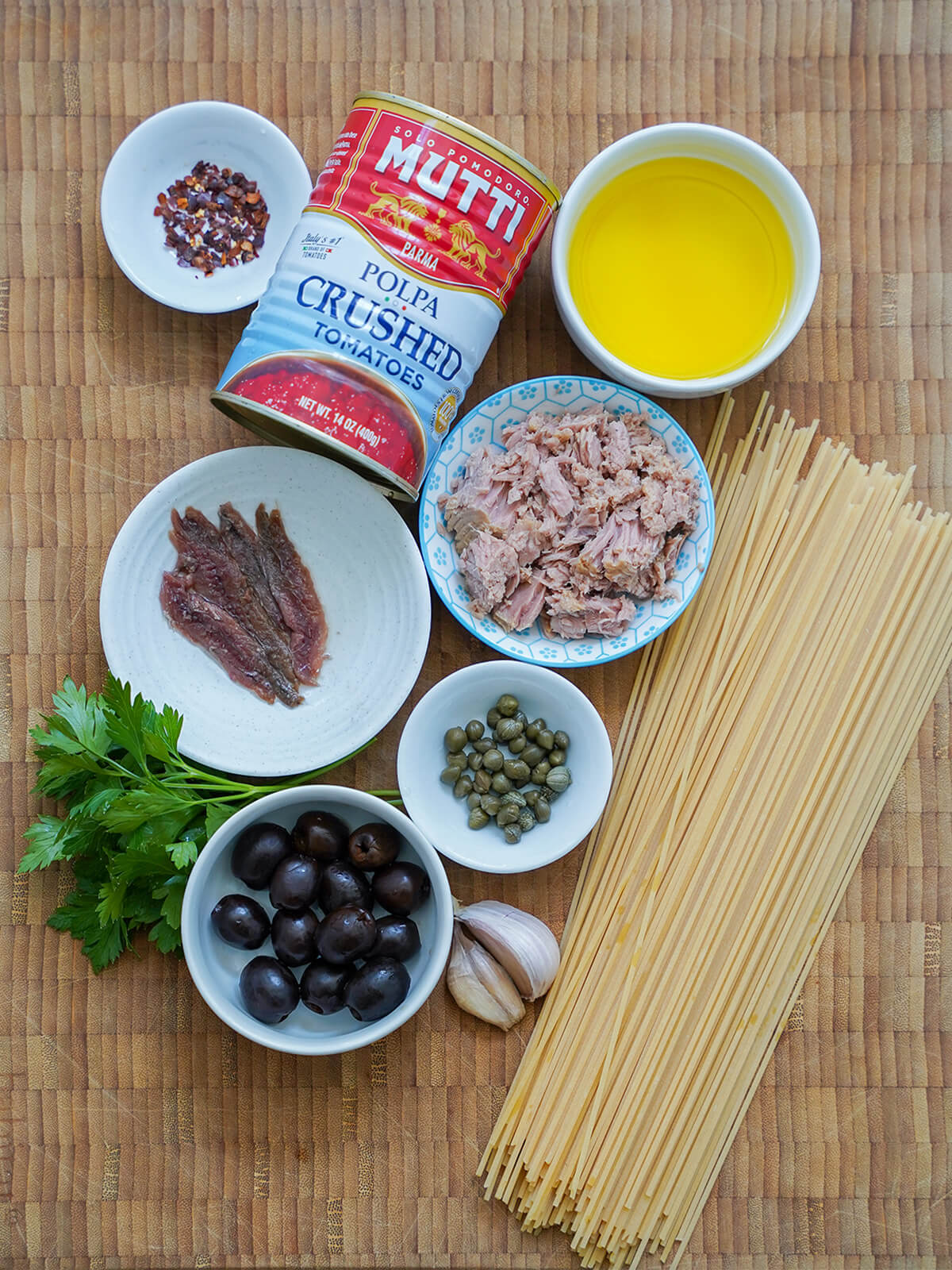 ingredients for tuna puttanesca can of tomatoes, spaghetti with dishes of oil, tuna, anchovies, capers, olives and red pepper flakes in between and some garlic and parsley to side.