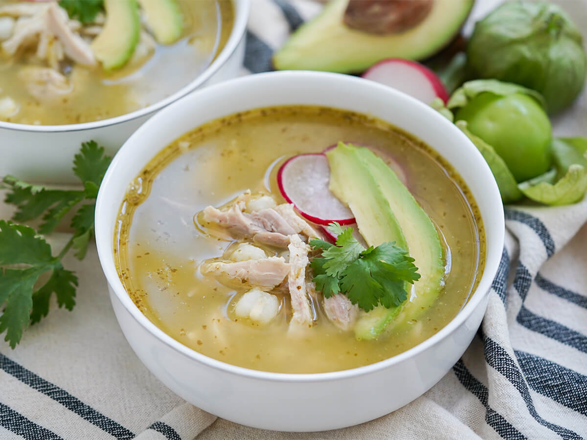 side view of bowl of pozole verde with tomatillos and part avocado behind.