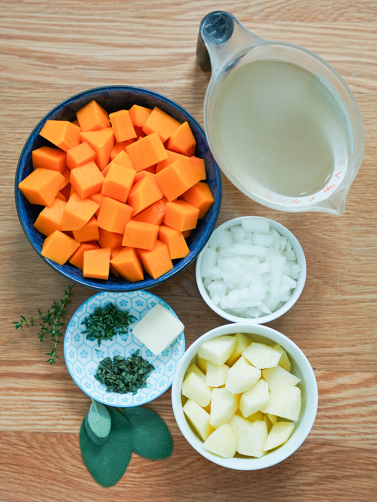 butternut squash soup ingredients bowls with chunks of squash, apple, onion, measure of stock, small dish with chopped herbs and butter.