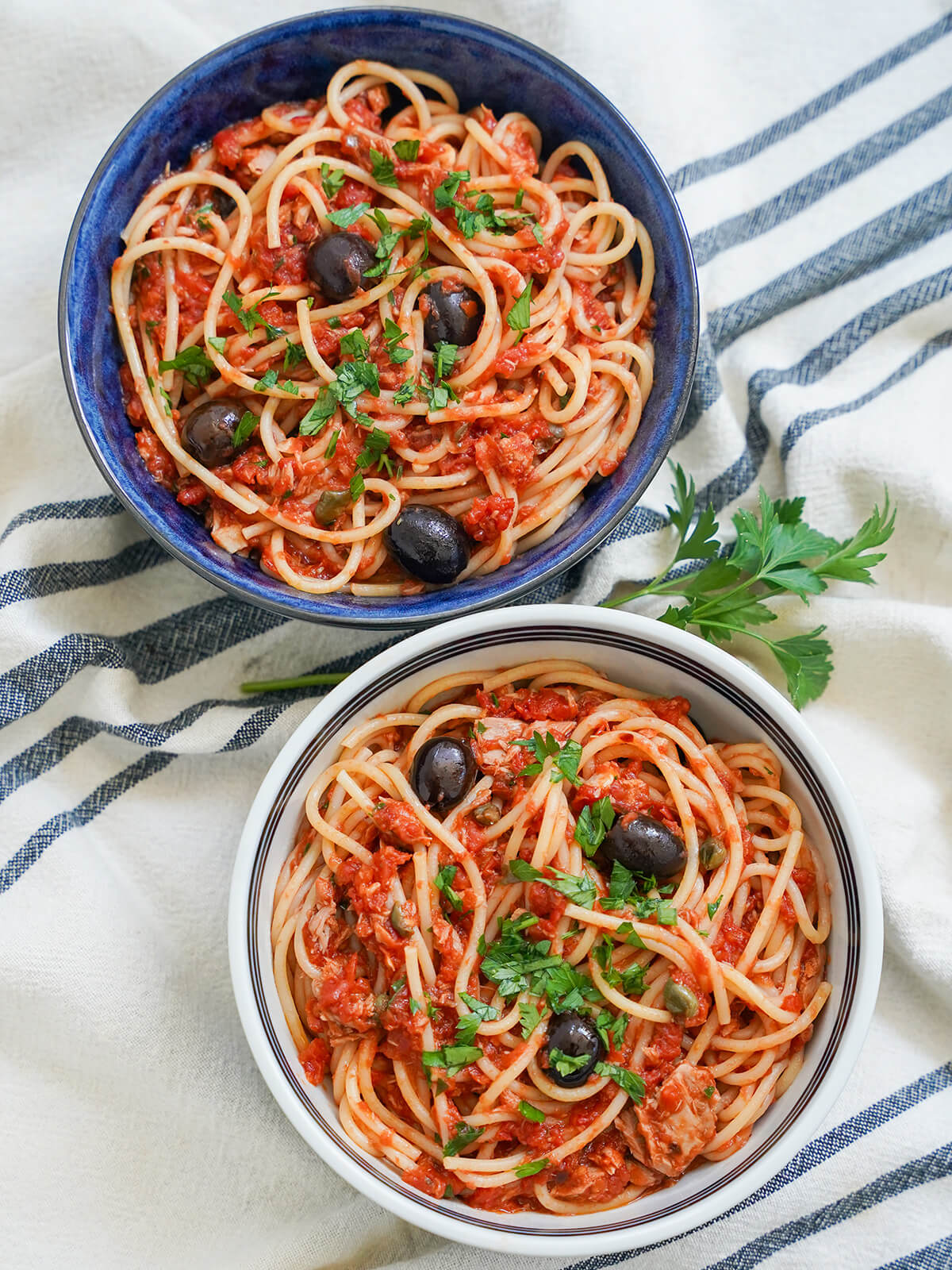 Pasta puttanesca might not be as well known as some other Italian dishes, but it really should be. A little saltiness from anchovies and capers, a little heat from red pepper, fresh tomatoes and meaty tuna. And incredibly easy to make. two bowls of tuna puttanesca with sprig of parsley between from overhead.