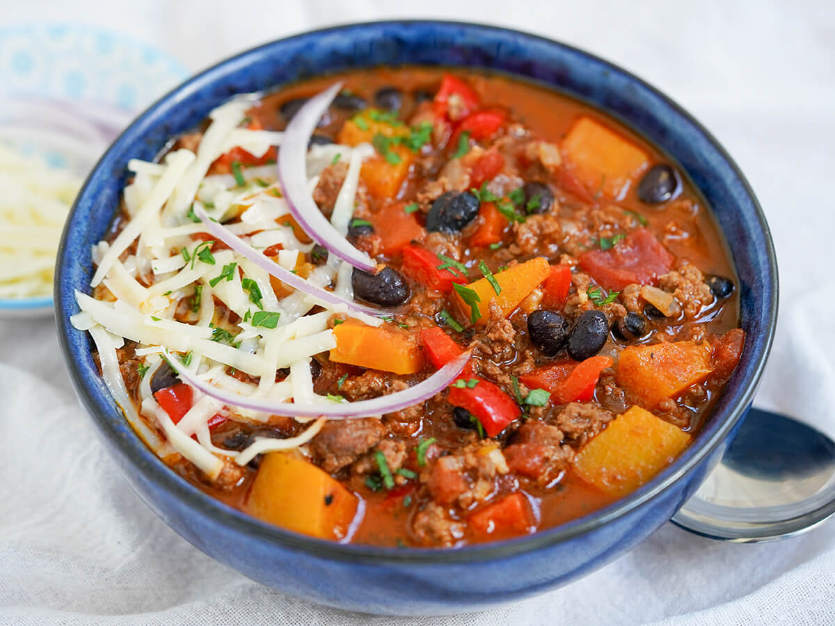 bowl of chili with butternut squash topped with cheese and red onion.