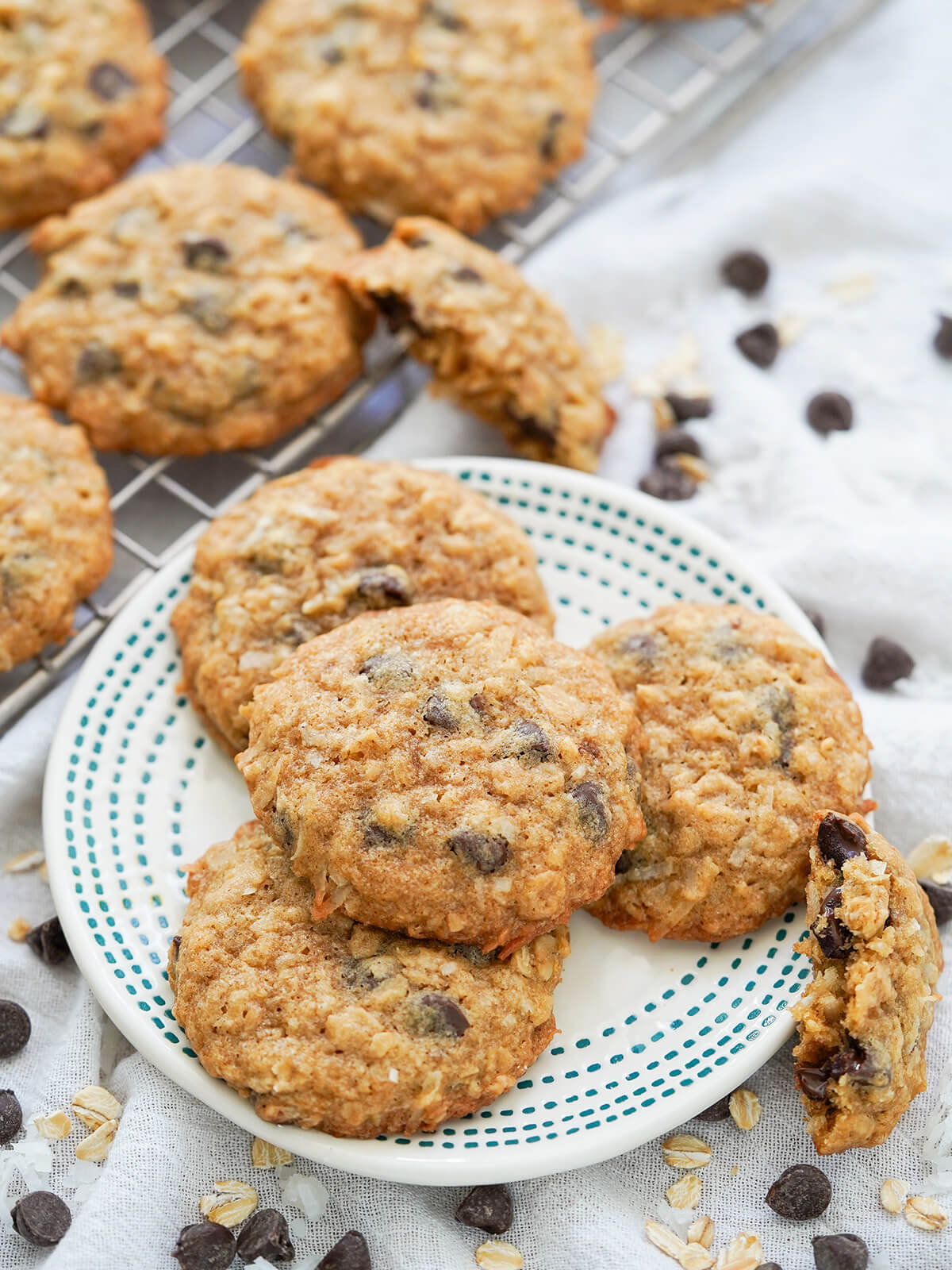 plate of coconut oatmeal chocolate chip cookies with more on rack behind and chocolate chips and oats scattered around plate.