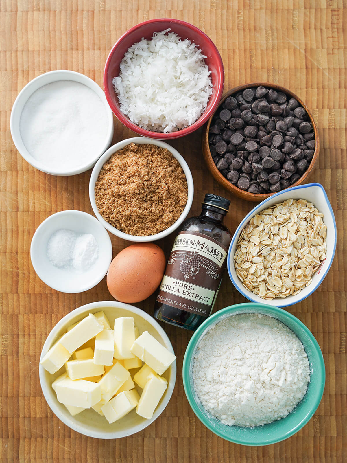 bowls of sugars, chocolate chips, shredded coconut, oats, flour, butter pieces and egg and vanilla bottle in middle.