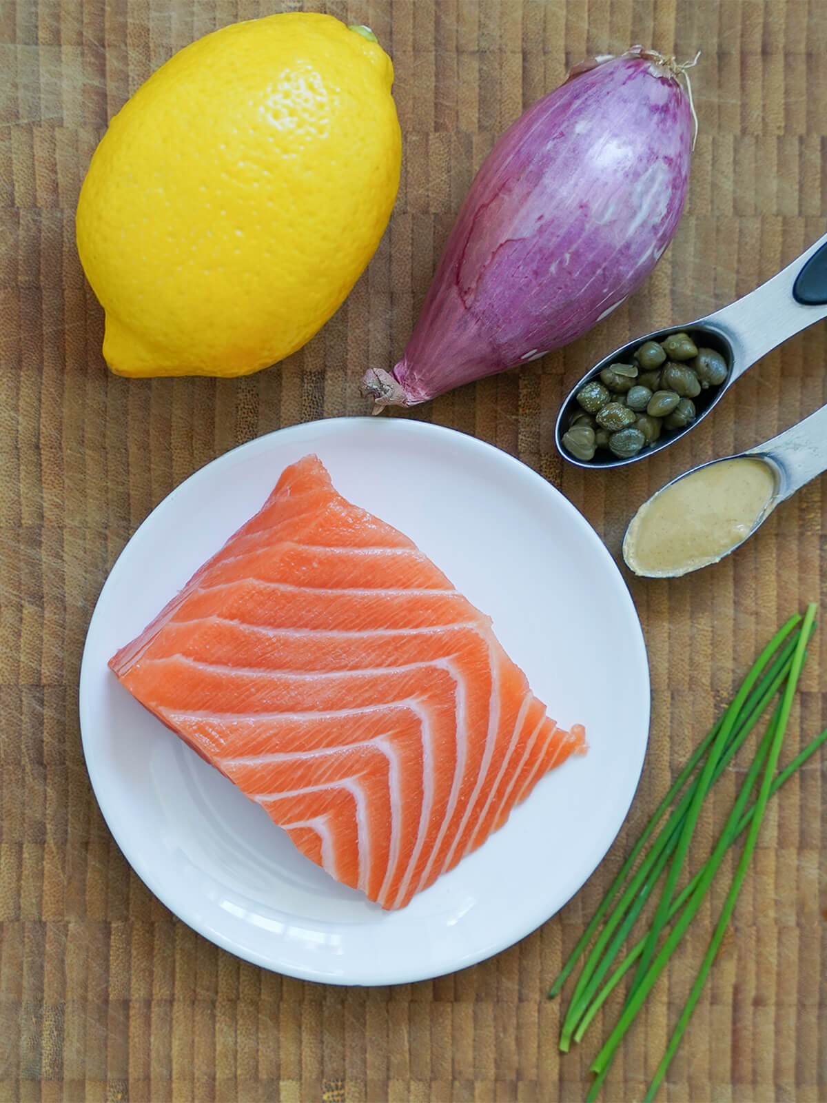 piece of salmon on plate with lemon, shallot, chives and spoons with capres and mustard around.