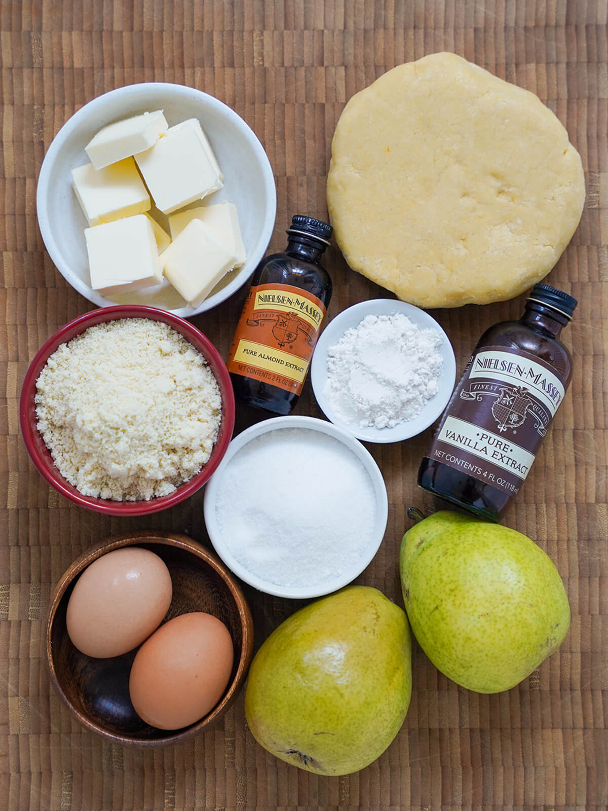 ingredients for pear frangipane tart dish with cubes of butter, bowls of almond flour, sugar, eggs, pears, piece of pastry dough and small bottles of extract on board.