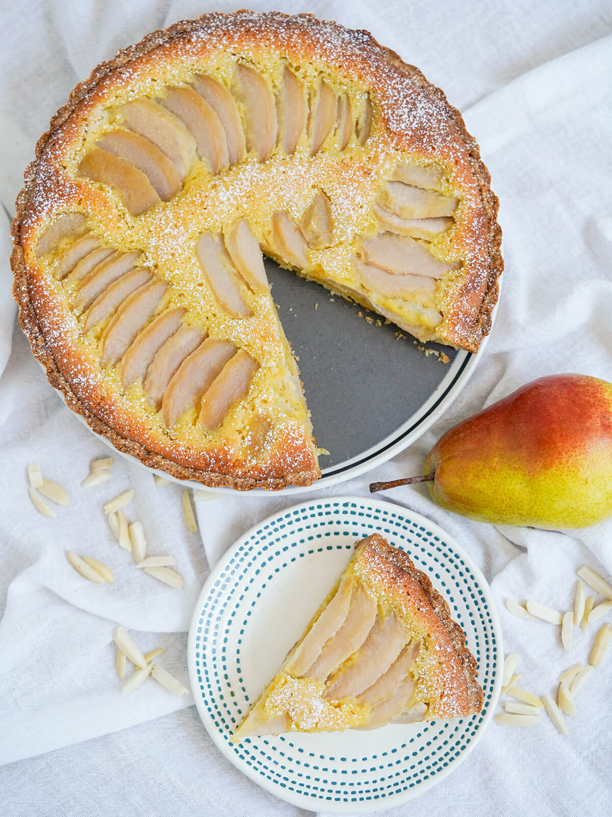 This pear frangipane tart is a delicious combination of tender pears nestled in a soft nutty filling, encased in crisp pastry. Elegant, shareable and perfect to enjoy any excuse. overhead view of pear frangipane tart with a slice cut out and the slice on plate below with pear to one side.