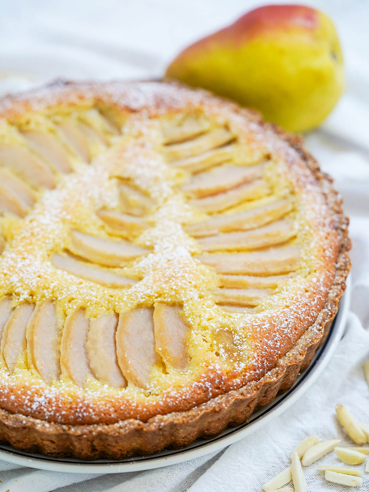 This pear frangipane tart is a delicious combination of tender pears nestled in a soft nutty filling, encased in crisp pastry. Elegant, shareable and perfect to enjoy any excuse. side view of pear frangipane tart with a pear behind.