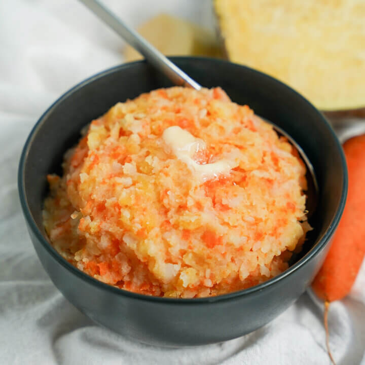 bowl of rotmos, Swedish root vegetable mash, with a spoon to back and vegetables in background.
