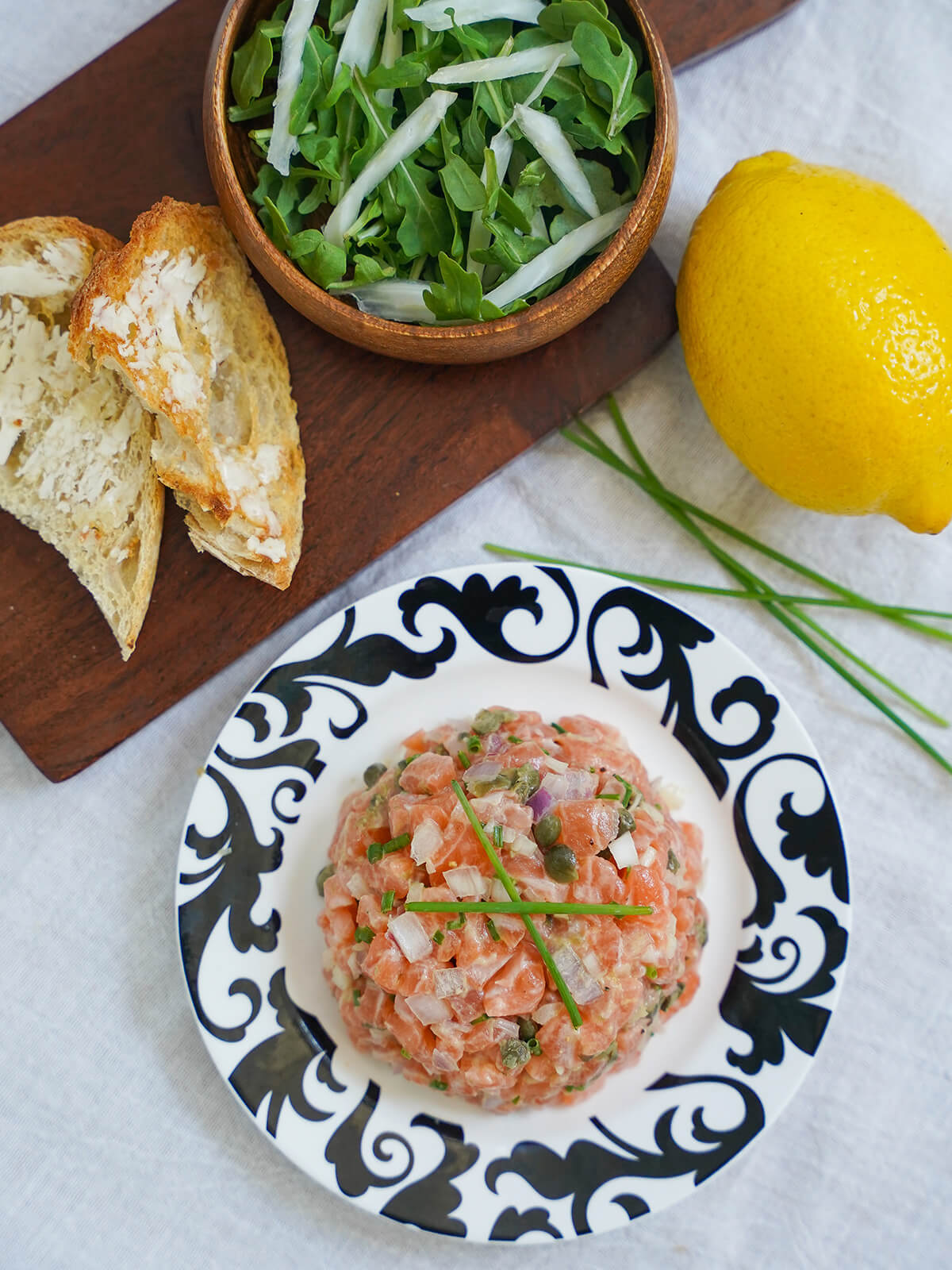 overhead view of salmon tartare on plate with side salad and buttered toast on board above, lemon to side.