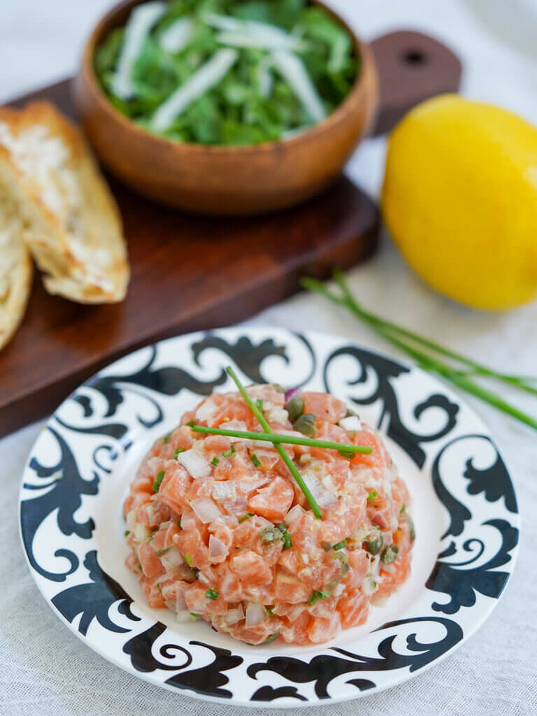 plate of salmon tartare garnished with chives with small salad, toast and lemon behind.