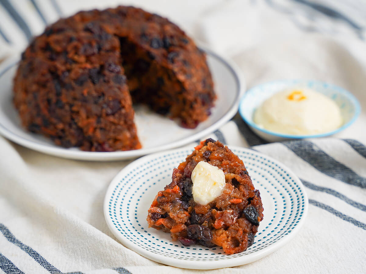 slice of Christmas pudding topped with brandy butter with rest of pudding on plate behind and more brandy butter to side.