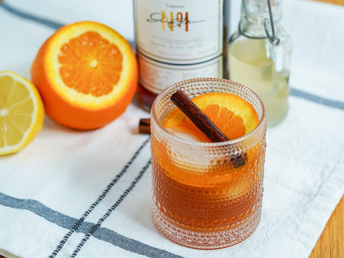 glass of Winter Pimms cup with cut orange and lemon behind, along with part view of bottles.