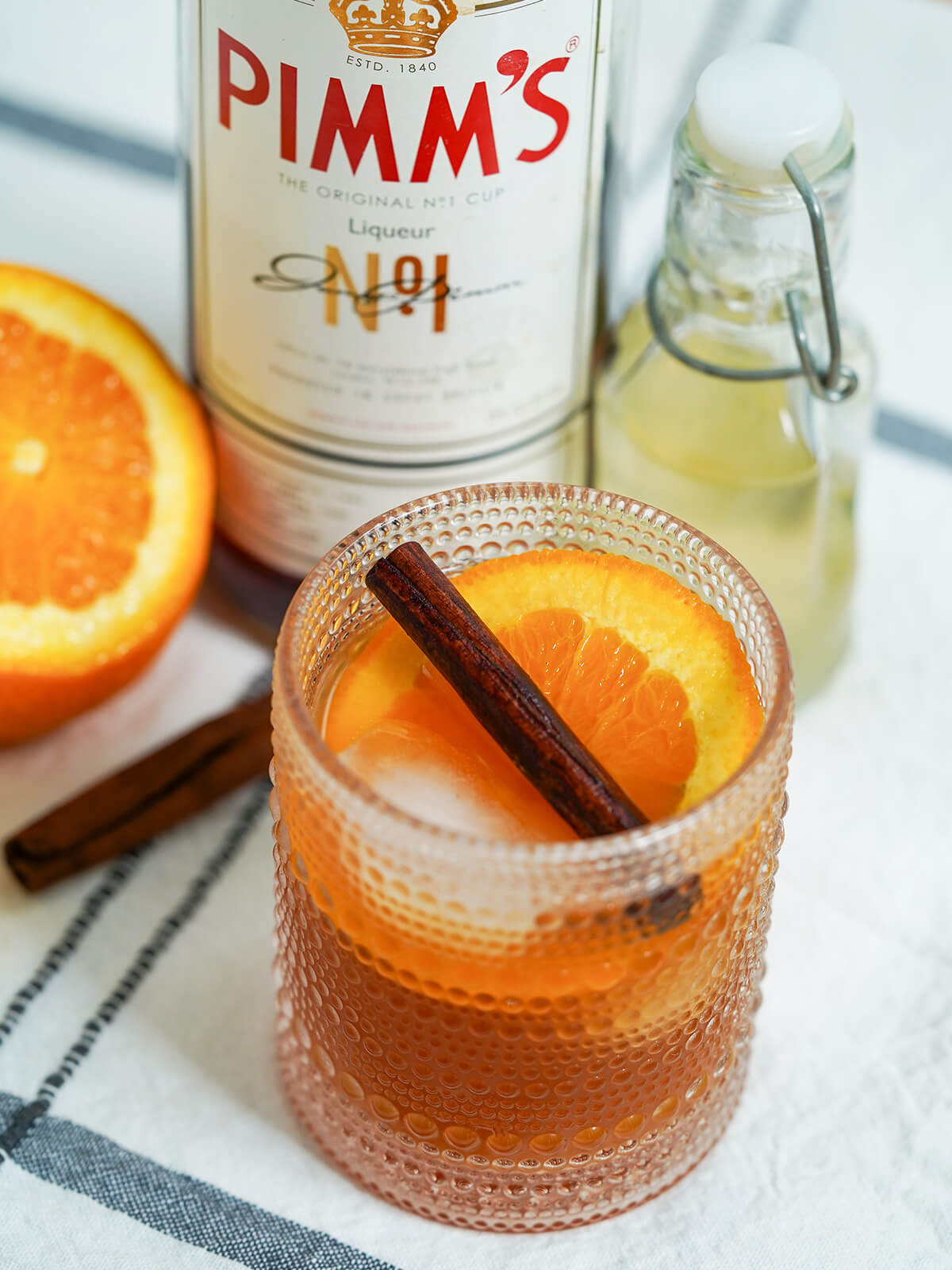 partly overhead view of Winter Pimms cup garnished with orange and cinnamon stick with bottle of Pimm's behind.
